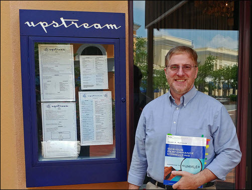 Photograph of Gary DePaul in front of a restaurant holding the book, Serious Performance Consulting